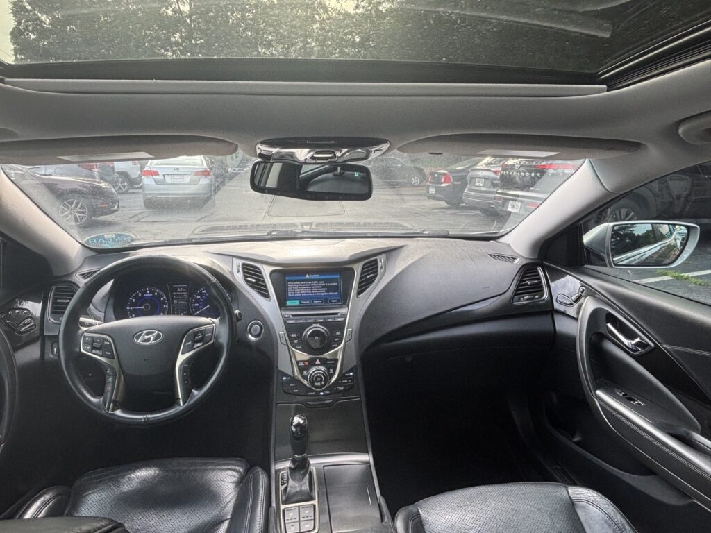 Interior view of a modern car cabin seen from the rear seats, showing the steering wheel, center console, and sunroof with parked cars outside the windshield.