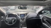 Interior view of a modern car cabin seen from the rear seats, showing the steering wheel, center console, and sunroof with parked cars outside the windshield.