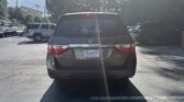 Rear view of a dark gray Honda Odyssey minivan parked in a dealership lot, with other cars in nearby spaces and sun glare on the rear window.
