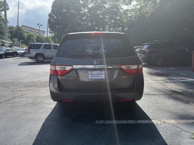 Rear view of a dark gray Honda Odyssey minivan parked in a dealership lot, with other cars in nearby spaces and sun glare on the rear window.