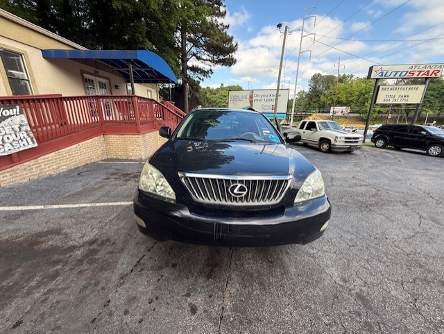 Front view of a black Lexus SUV parked in a used-car lot, with a brick building and red railing on the left under a blue awning.