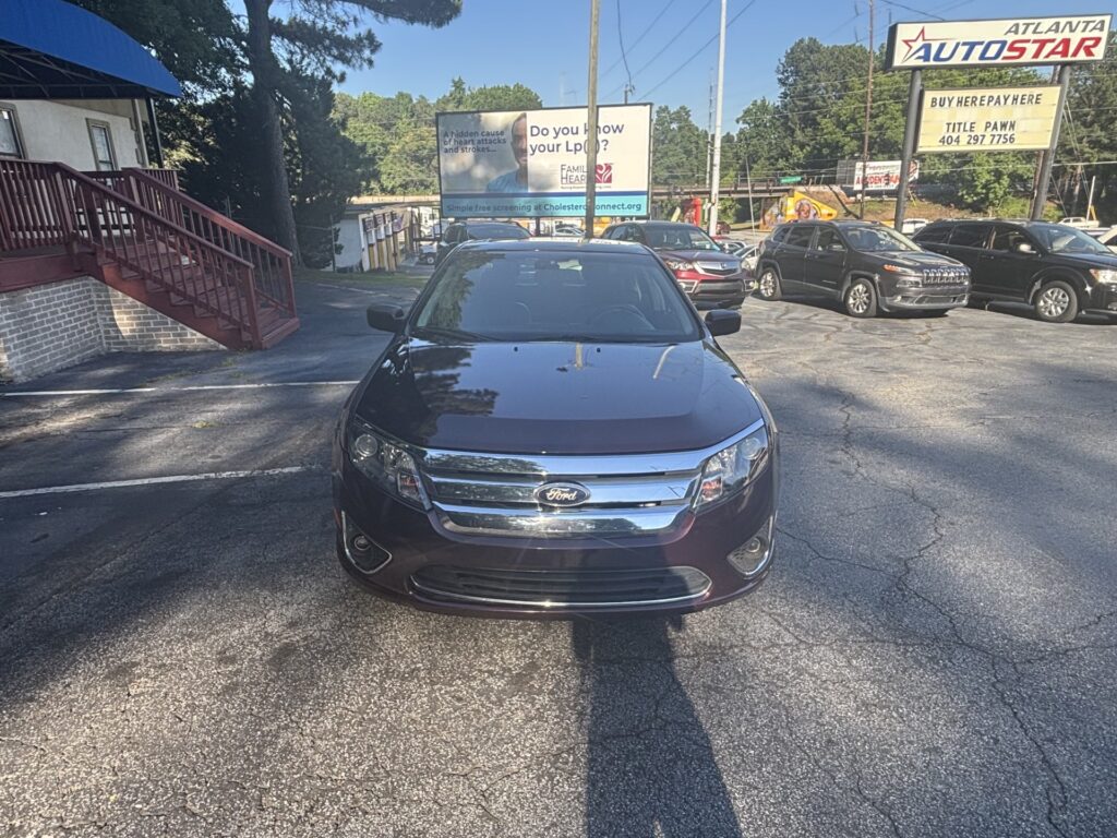 Front view of a dark Ford sedan parked in a lot, with a large roadside billboard and other vehicles in the background.