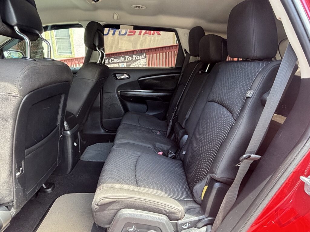 Rear row of a red car with black fabric seats, three seating positions, headrests, and visible seat belts from the open door.