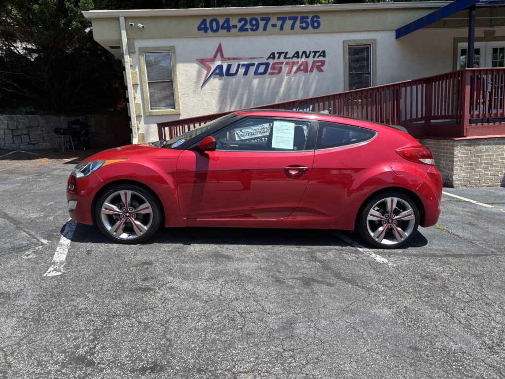 Red hatchback car parked in a lot in front of an Atlanta AutoStar dealership building with blue signage above the entrance area and a wooden ramp to the right.