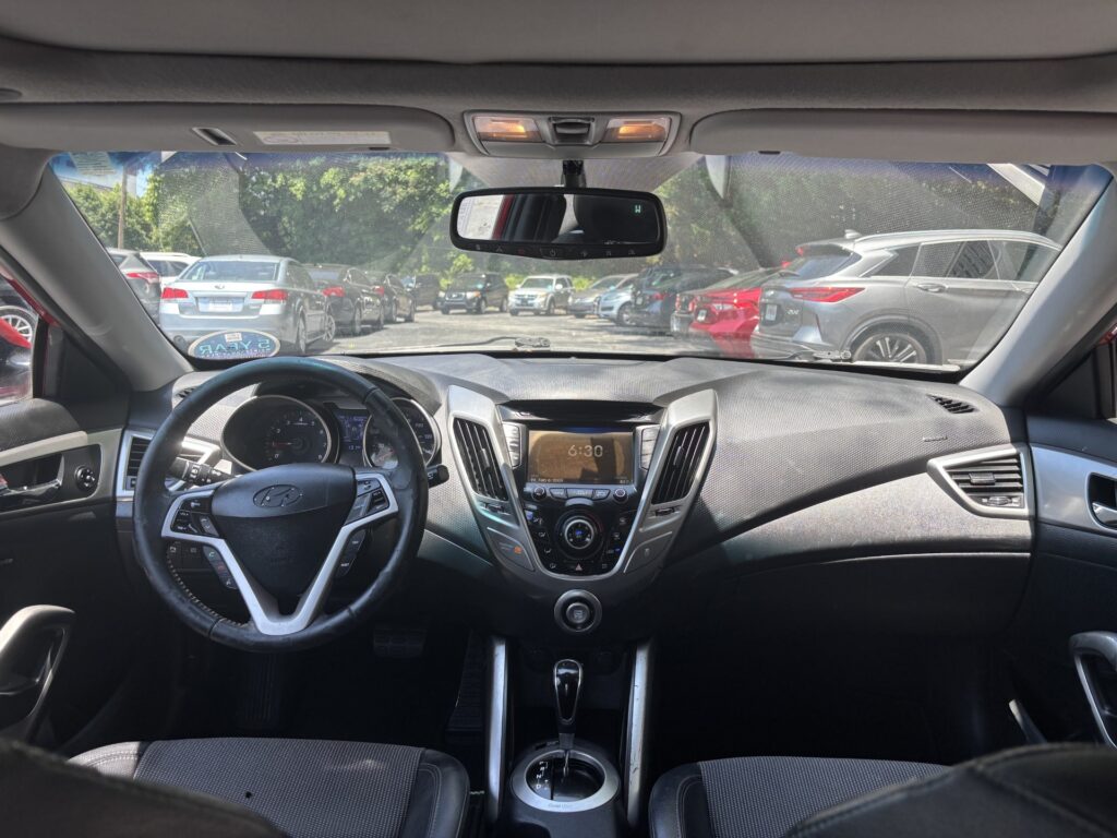 View of a car's interior from the back seat, showing the steering wheel, center console, and dashboard with outside parked cars visible through the windshield/side windows.