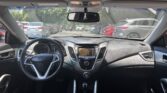 View of a car's interior from the back seat, showing the steering wheel, center console, and dashboard with outside parked cars visible through the windshield/side windows.