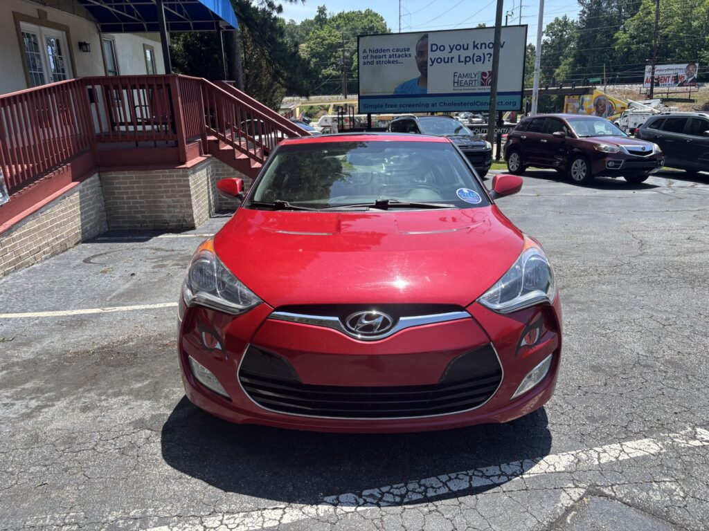 Front view of a red Hyundai sedan in a parking lot with a brick building and staircase on the left and a large billboard in the background.
