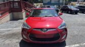Front view of a red Hyundai sedan in a parking lot with a brick building and staircase on the left and a large billboard in the background.
