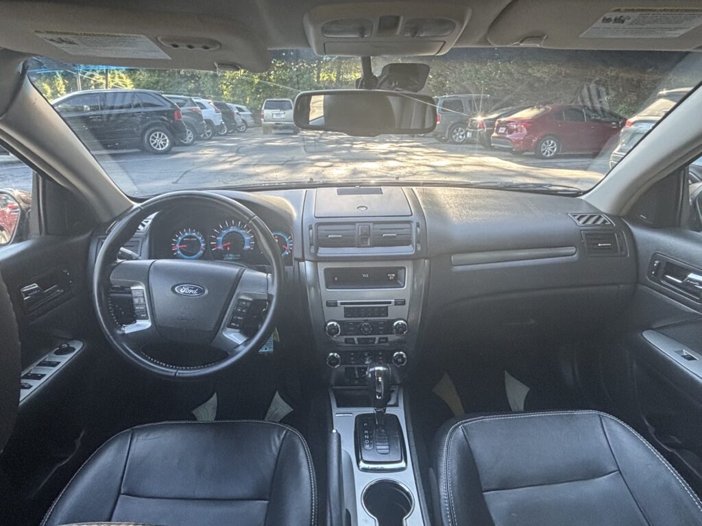 Driver's view of a car interior: steering wheel, center console, and gear shifter, with a row of parked cars visible through the windshield.