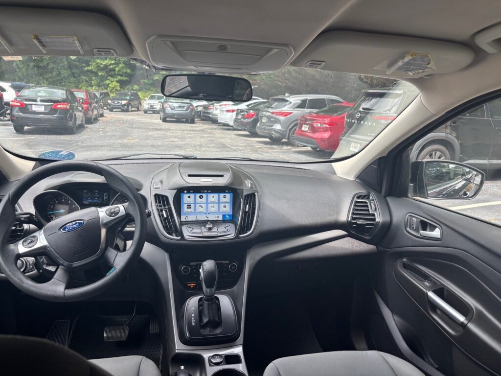 Driver's-seat view of a Ford interior with steering wheel, center touchscreen, and gear shifter, parked in a lot with other cars outside.