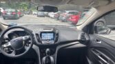 Driver's-seat view of a Ford interior with steering wheel, center touchscreen, and gear shifter, parked in a lot with other cars outside.